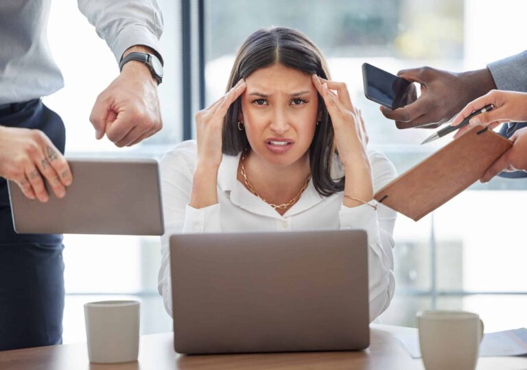 A stressed woman at a desk holds her head while surrounded by people's hands offering tablets, phones, and documents, emblematic of the ridiculous workplace rules overwhelming her daily life.
