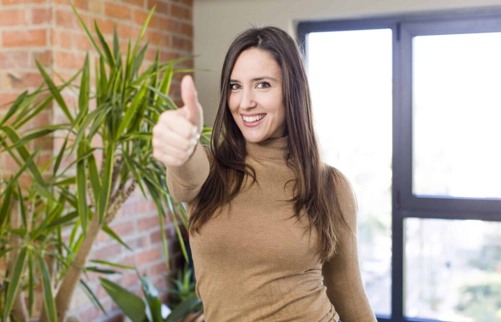 A woman in a brown turtleneck gives a thumbs-up in front of a plant and large windows.