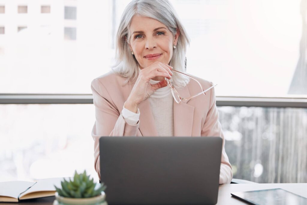 Older woman with gray hair in a peach blazer sits at a desk holding glasses, smiling at the camera. A laptop, plant, and notebook are on the desk in front of her.