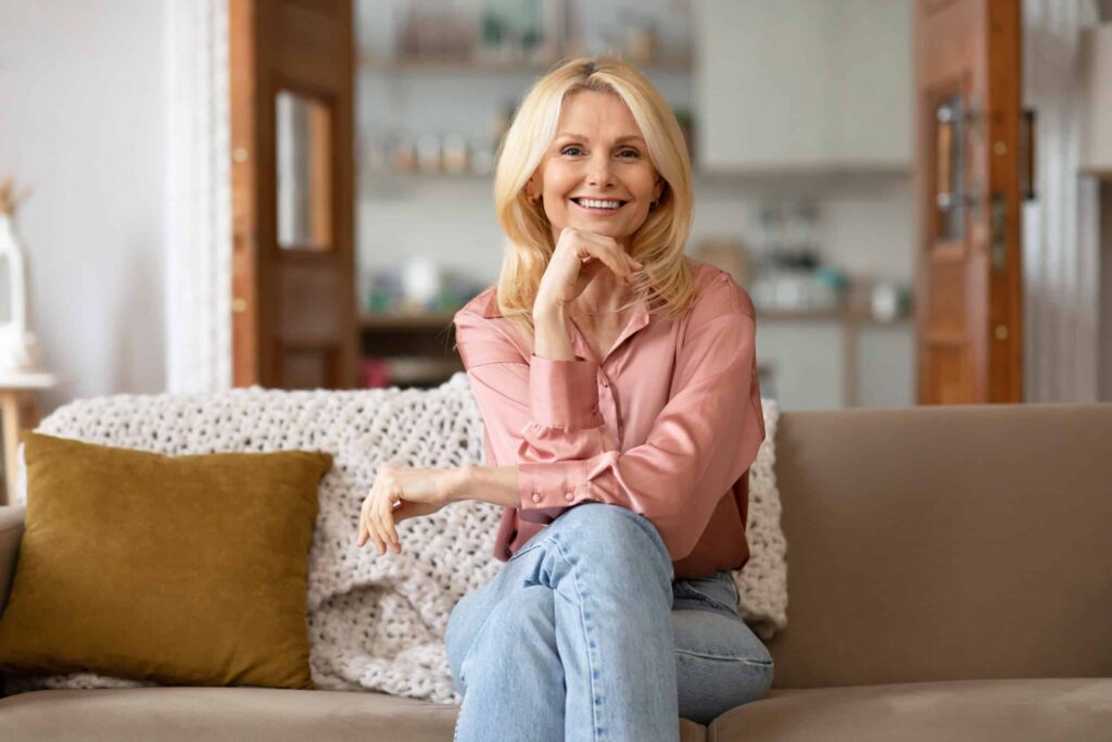 A woman with blonde hair sits on a sofa, wearing a pink blouse and blue jeans, smiling with her hand under her chin. The background features a cozy indoor setting.
