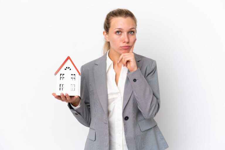 A woman in a gray suit holds a small model house, pondering the hidden costs of homebuying with a thoughtful expression.