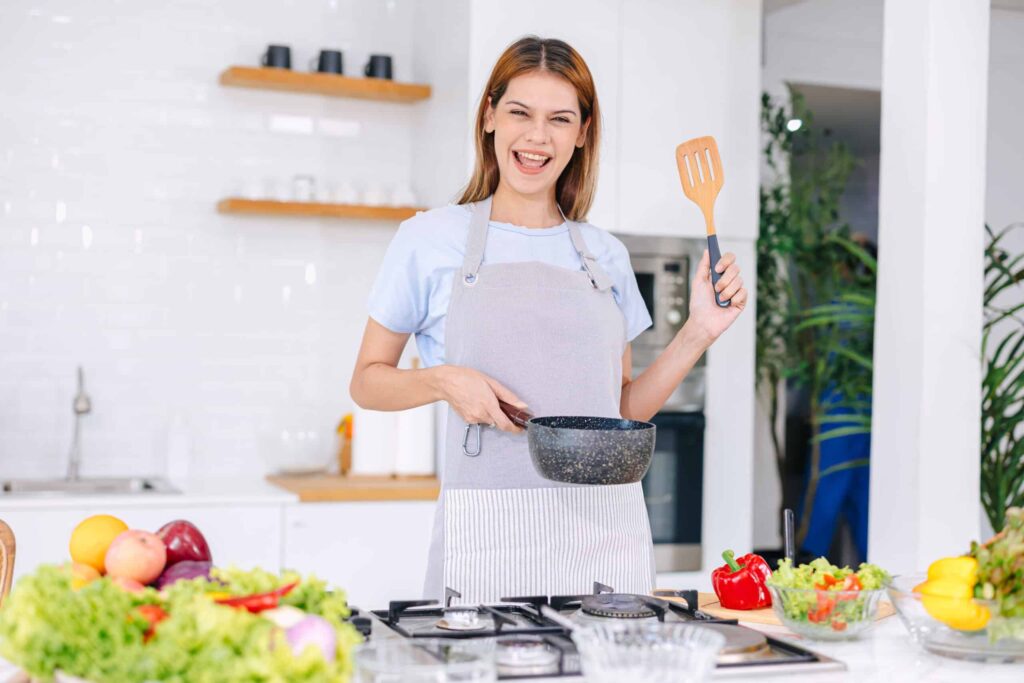Person wearing an apron smiling while holding a pot and spatula in a kitchen, surrounded by vegetables and fruits.