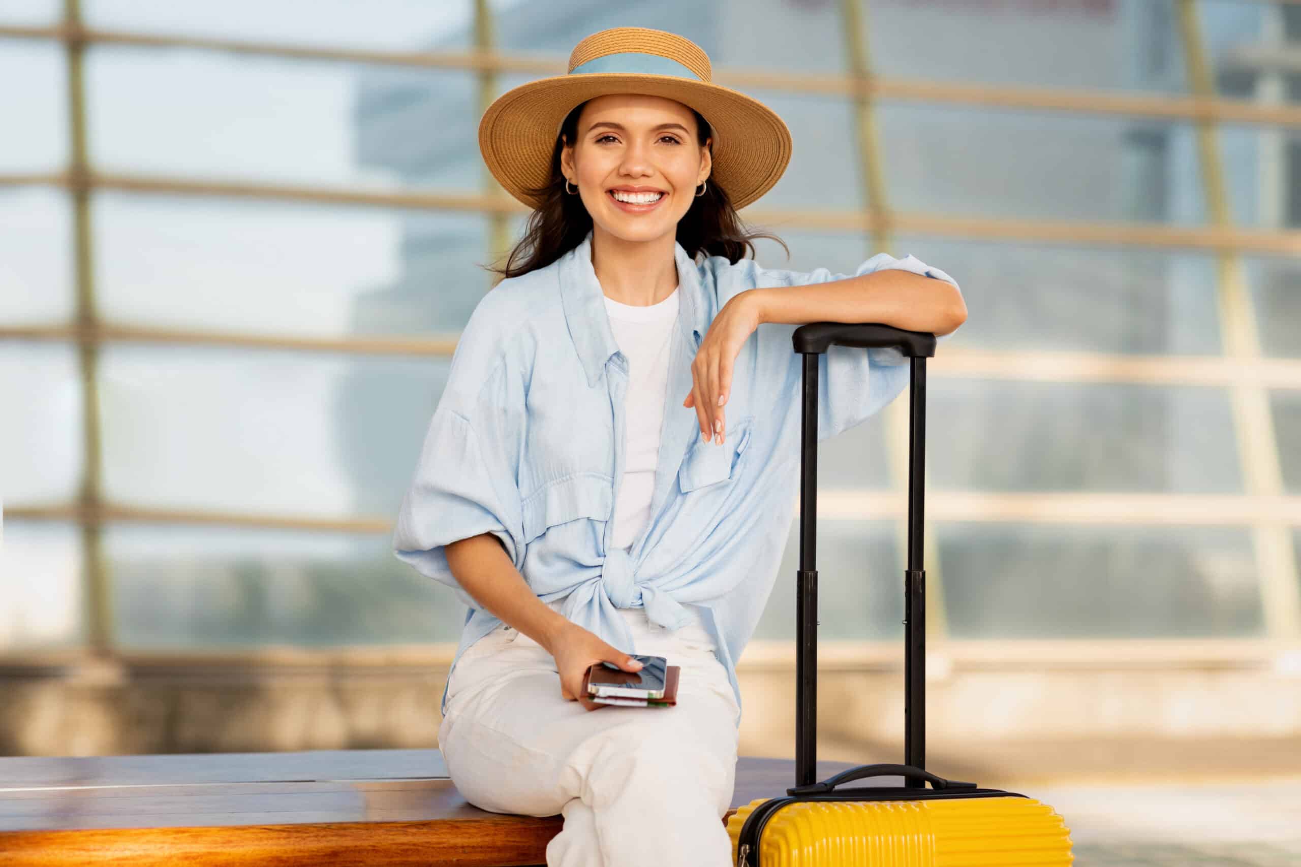 A woman in a straw hat and casual attire sits on a bench with a yellow suitcase and holds a phone and passport. She is smiling and appears relaxed.