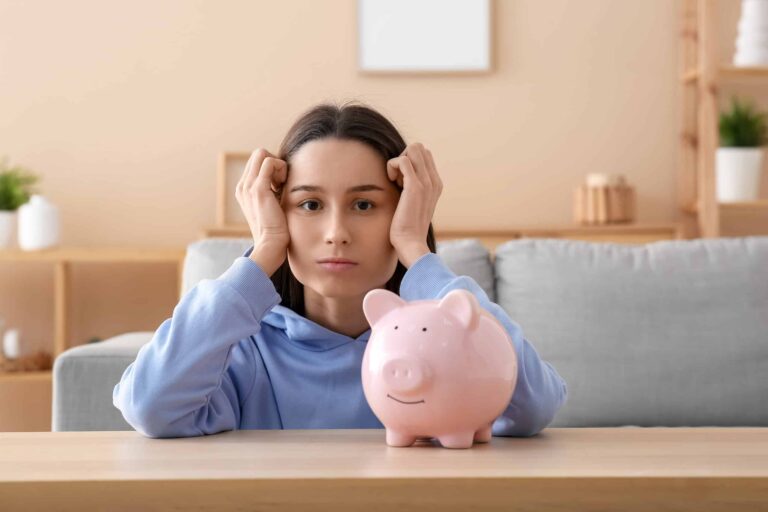 A woman in a blue hoodie sits at a table, resting her chin on her hands, looking at a pink piggy bank. A sofa is in the background.