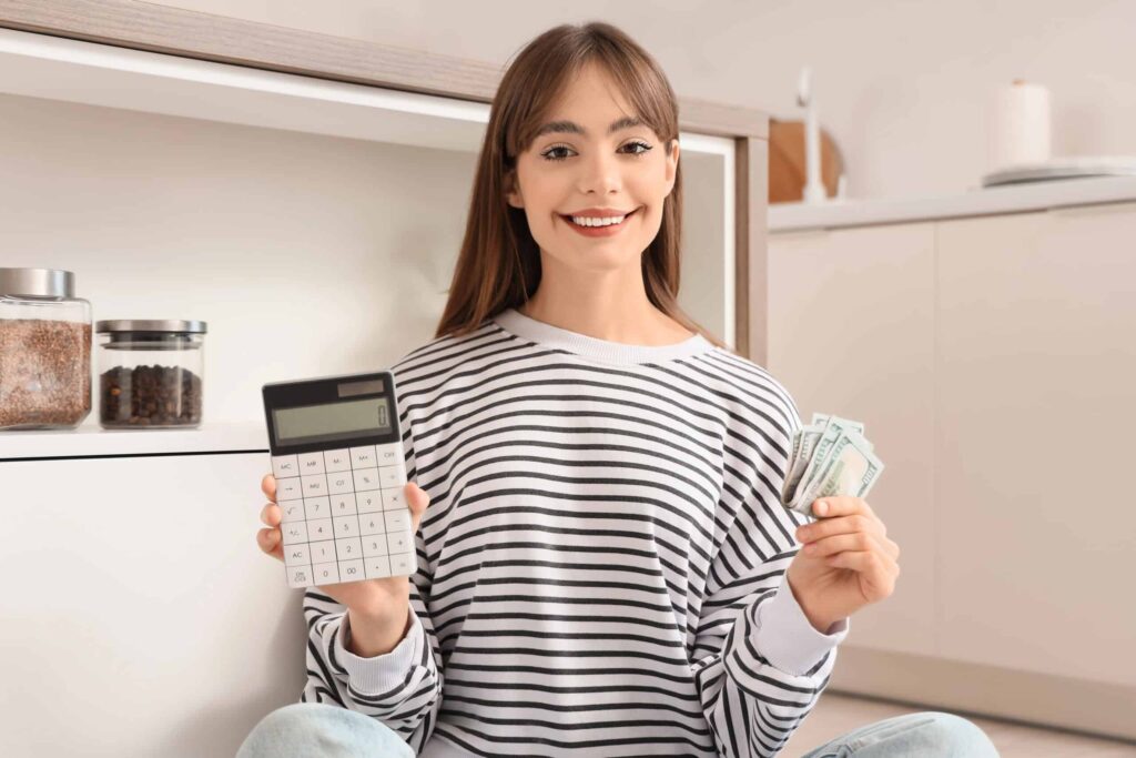Woman in a striped shirt sitting on the floor, holding a calculator in one hand and dollar bills in the other, smiling at the camera.