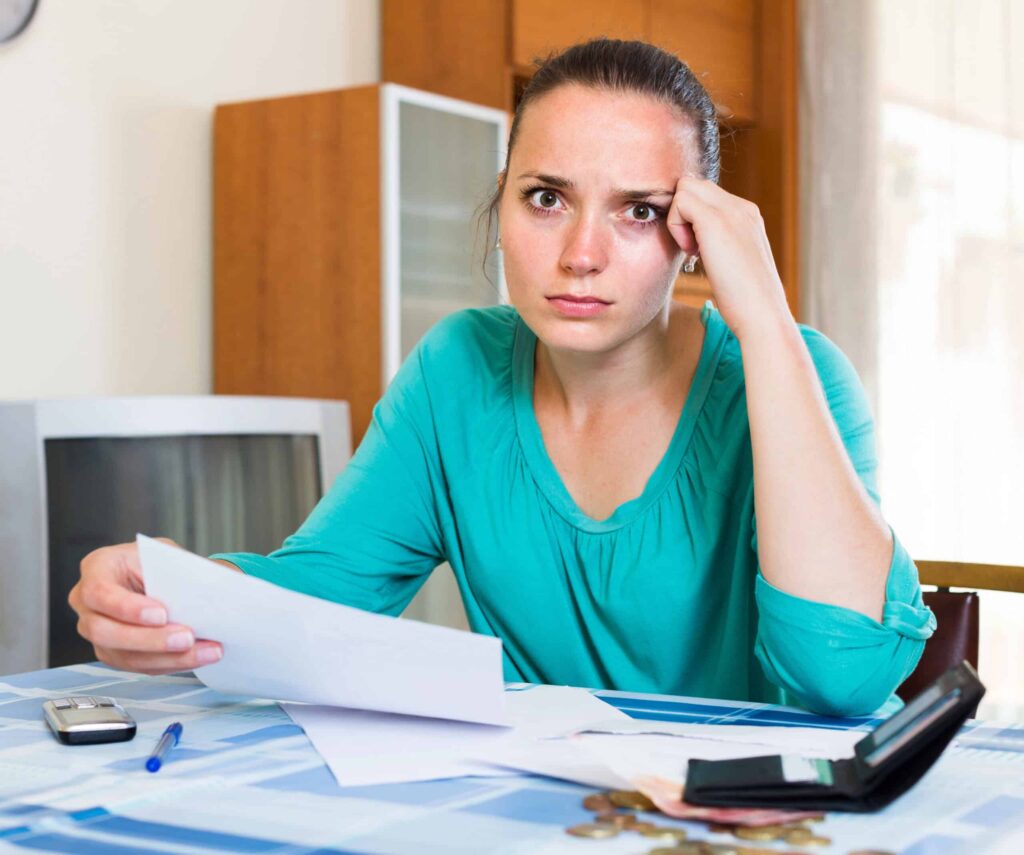 A woman sits at a table looking concerned, holding a piece of paper, with coins, a calculator, and a pen nearby.