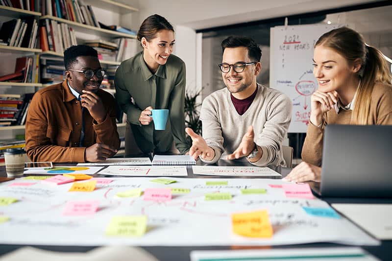 Four people in a workspace collaborate on a project, surrounded by colorful sticky notes and charts. One person gestures while speaking; another holds a mug. A laptop is on the table.