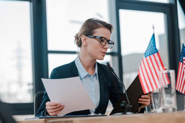 A woman in glasses speaks at a microphone, holding papers. Two small U.S. flags are on the table in front of her.