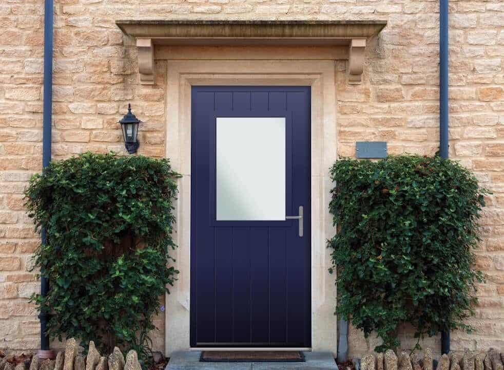 A navy blue door with a rectangular window is set in a stone wall. Two neatly trimmed bushes flank the entrance, and a black outdoor lamp is mounted on the left side.