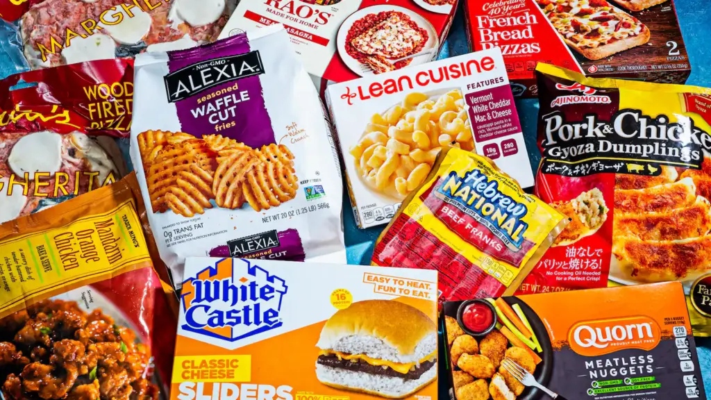 A colorful assortment of packaged frozen foods, including pizza, fries, dumplings, pasta, sliders, chicken nuggets, and more, arranged on a blue surface.