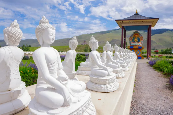 A row of white Buddha statues sits on stone platforms in a garden, with a colorful shrine in the background and rolling hills under a cloudy sky.