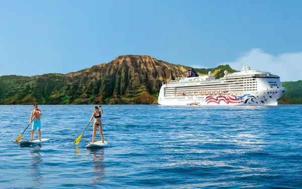 Two people paddleboarding on a calm ocean with a cruise ship in the background, set against a backdrop of rugged hills under a clear blue sky.