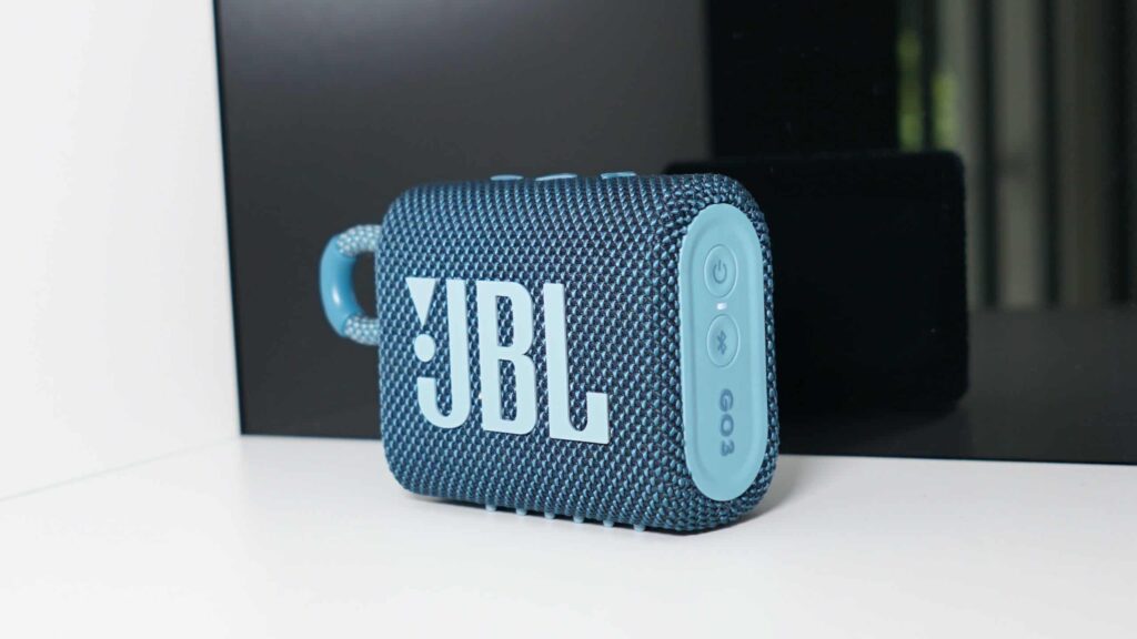 A blue JBL portable speaker with fabric texture is positioned upright on a white surface against a black background.