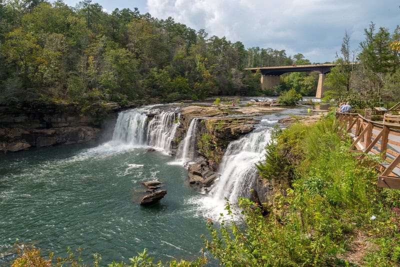 Waterfall cascading over rocks into a river, surrounded by trees, with a bridge and people visible in the background.