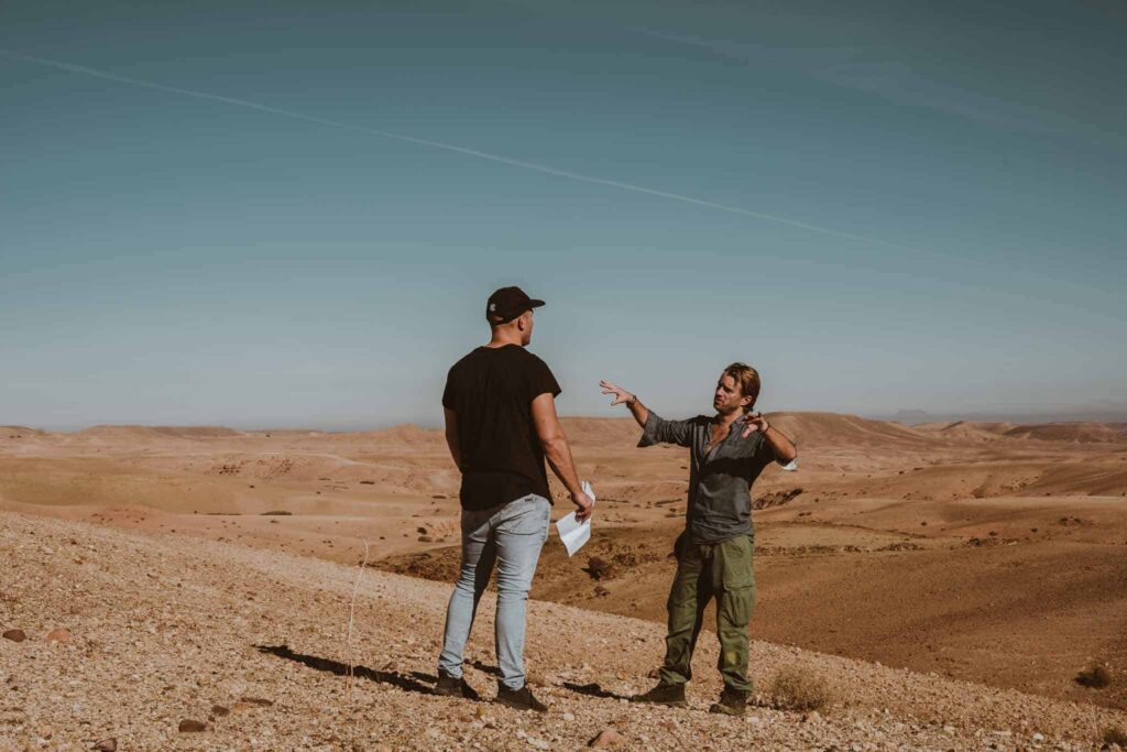 Two people stand and gesture to one another in a dry, expansive desert landscape under a clear blue sky.