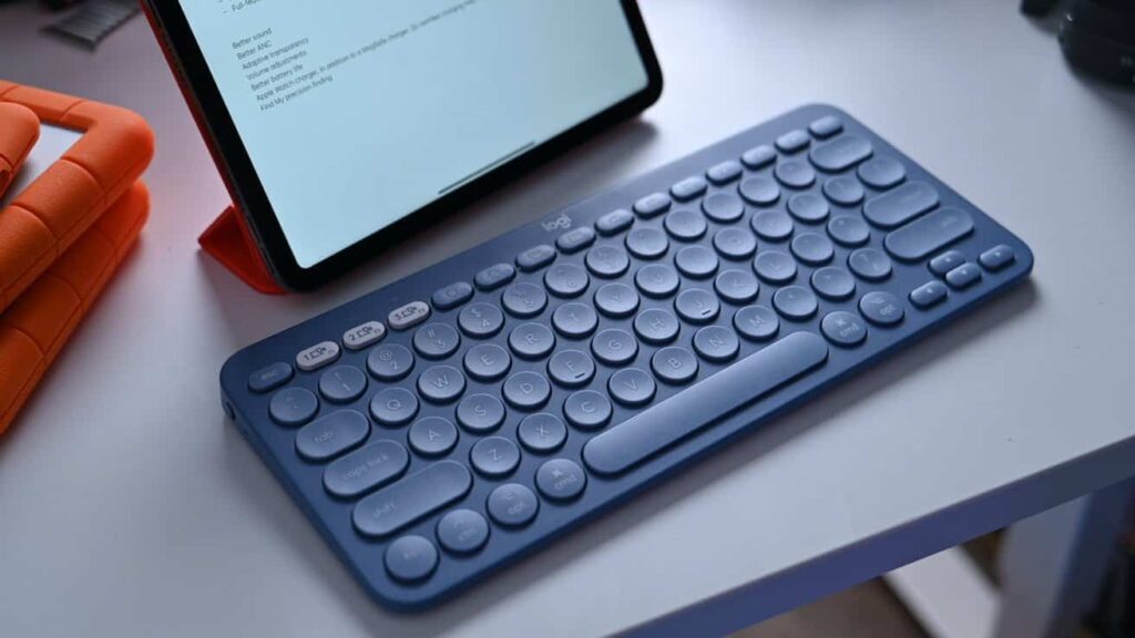 A blue wireless keyboard sits on a desk beside a tablet displaying text. An orange case is partially visible on the left.