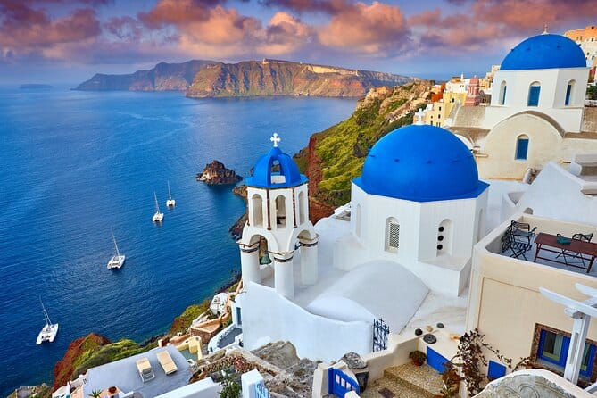 A scenic view of Santorini, featuring white buildings with blue domes overlooking the Aegean Sea, with sailboats and a rocky coastline in the background.
