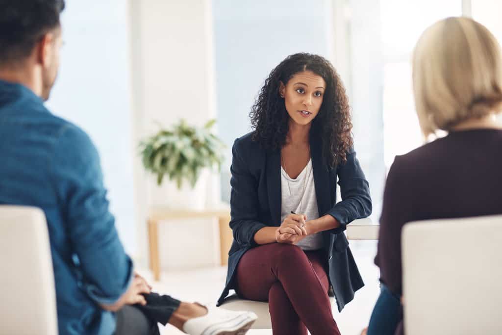 A woman with curly hair sits and talks to two people in a bright room with a potted plant in the background.