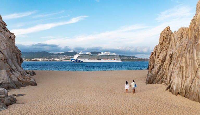 A cruise ship sails near a rocky beach with two people walking on the sand, clear blue sky above.