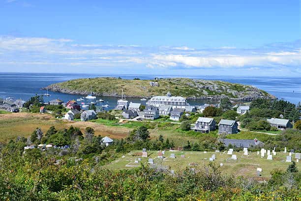 Scenic coastal village with houses, greenery, and a small bay. Rocky island and ocean visible in the background under a partly cloudy sky.