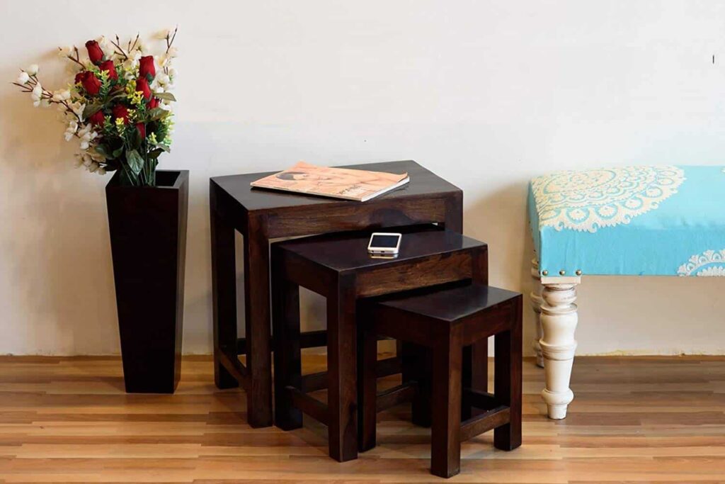 Nesting tables with a phone and magazine on top, next to a tall vase with red and white flowers and a blue cushioned bench on a wooden floor.