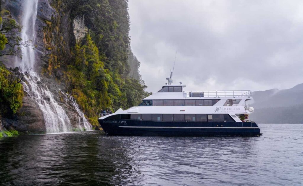 A white and blue tourist boat named "Fiordland Jewel" cruises near a rocky cliff and waterfall in a misty, forested area.