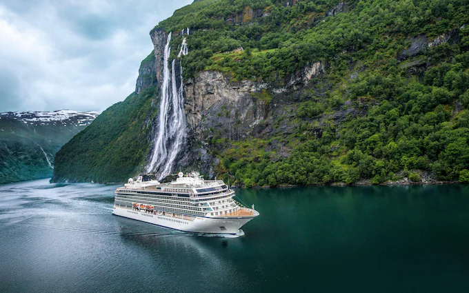 A cruise ship sails near a steep cliff with a waterfall cascading into a fjord surrounded by lush greenery and cloudy skies.