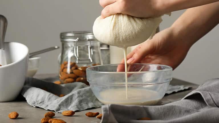 Hands squeezing almond milk from a cloth into a glass bowl, with almonds and a jar in the background.