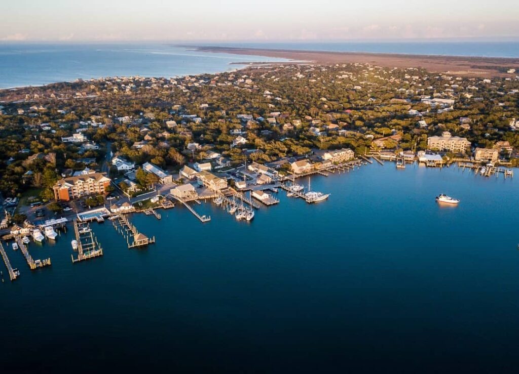 Aerial view of a coastal town with numerous docks and boats, surrounded by lush greenery and bordered by the ocean under a clear sky.