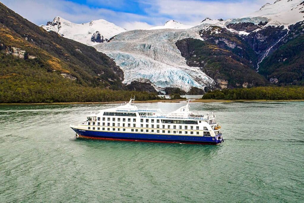 Cruise ship sailing near a glacier surrounded by snow-capped mountains and green forests.