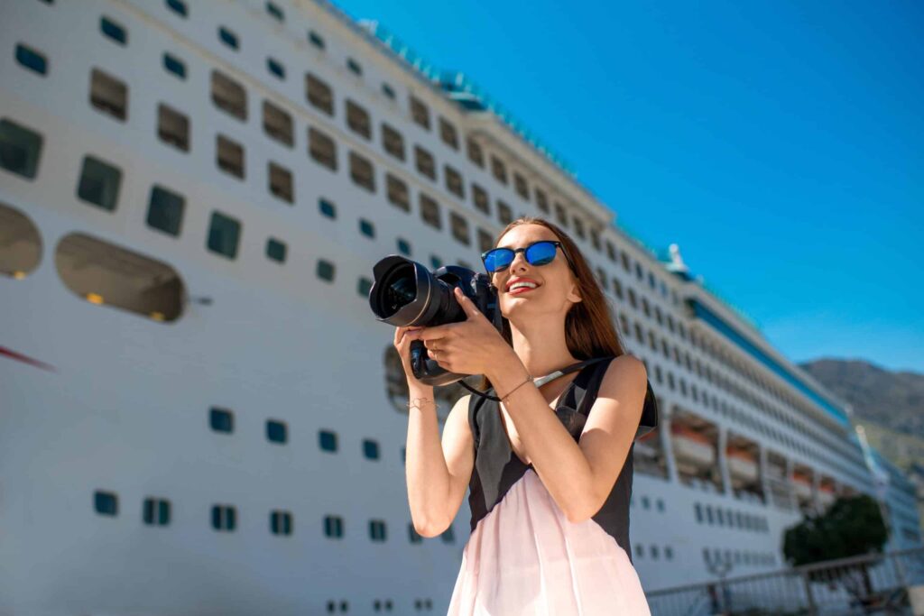 A woman with a camera smiles in front of a large cruise ship.