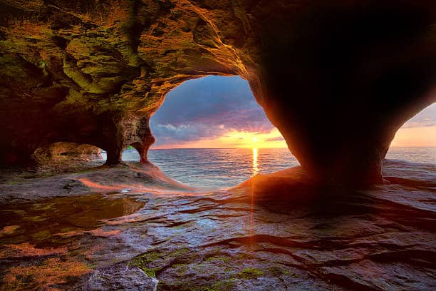 Sunset view from inside a rocky cave, with light streaming through the arched opening, reflecting on the water and stone floor.