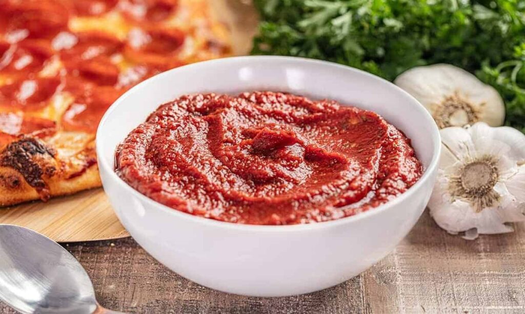 Bowl of tomato sauce on a table with pizza, fresh parsley, garlic, and a spoon nearby.