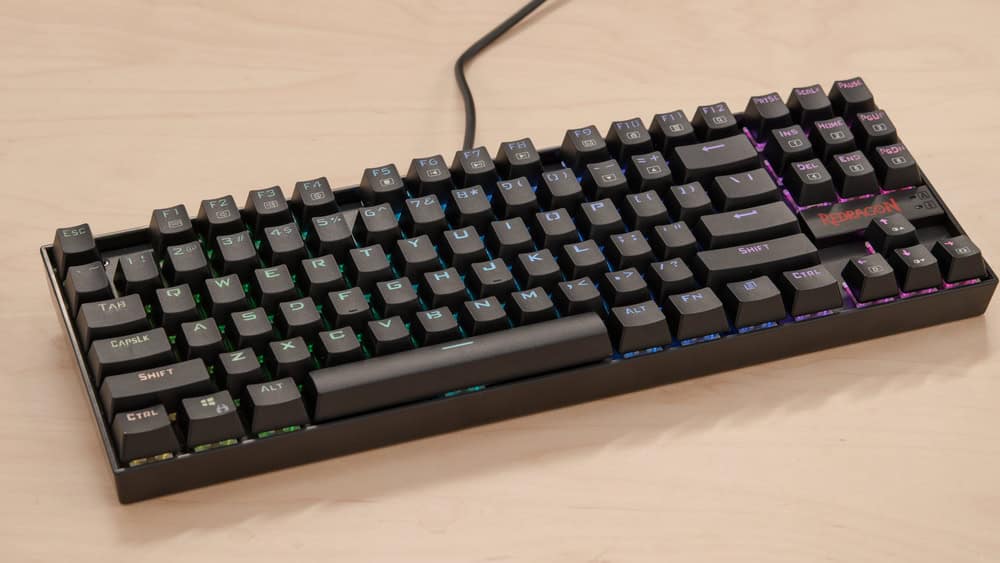 A black mechanical keyboard with rainbow backlit keys on a light wood surface.