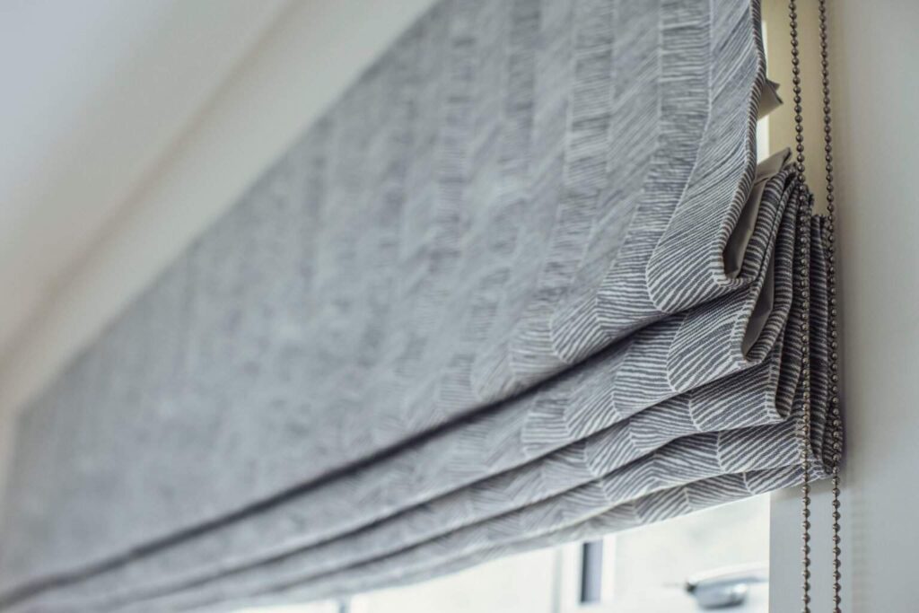 Close-up of a gray fabric Roman blind, partially raised, with a visible chain mechanism on the right side.