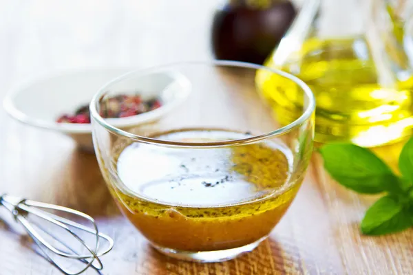 A glass bowl of vinaigrette dressing on a wooden surface, with a whisk and a small dish of spices in the background.