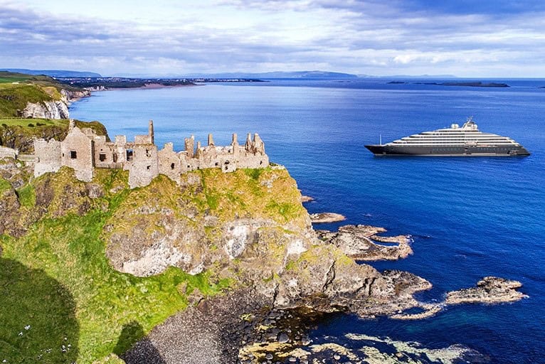 A large yacht sails near a rocky coastline with a historic stone castle ruin, under a lightly cloudy sky.