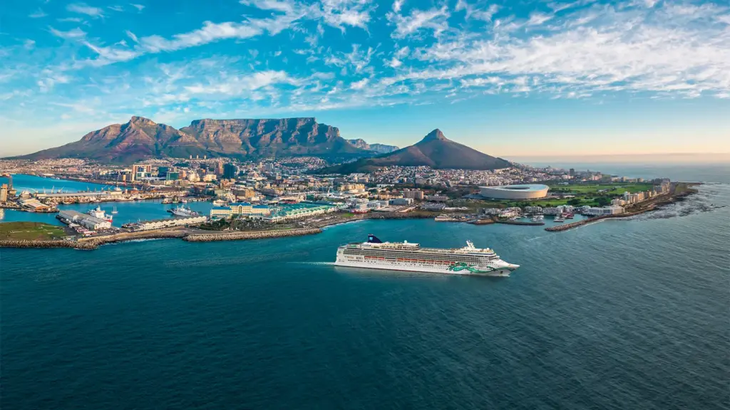Cruise ship sailing near a coastal city with a backdrop of a large, flat-topped mountain and clear blue sky.