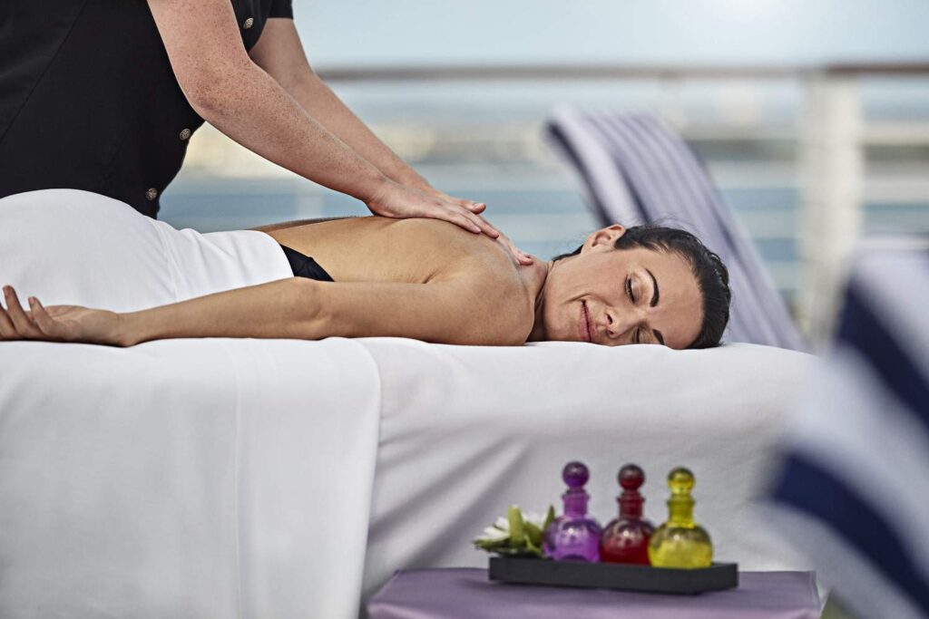 A person receives a back massage on a massage table outdoors, with colorful small bottles on a nearby tray.