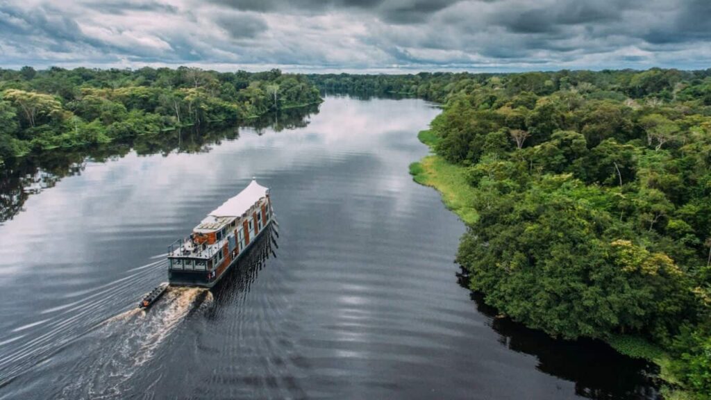 A boat navigates a wide, calm river bordered by lush green forest under a cloudy sky.