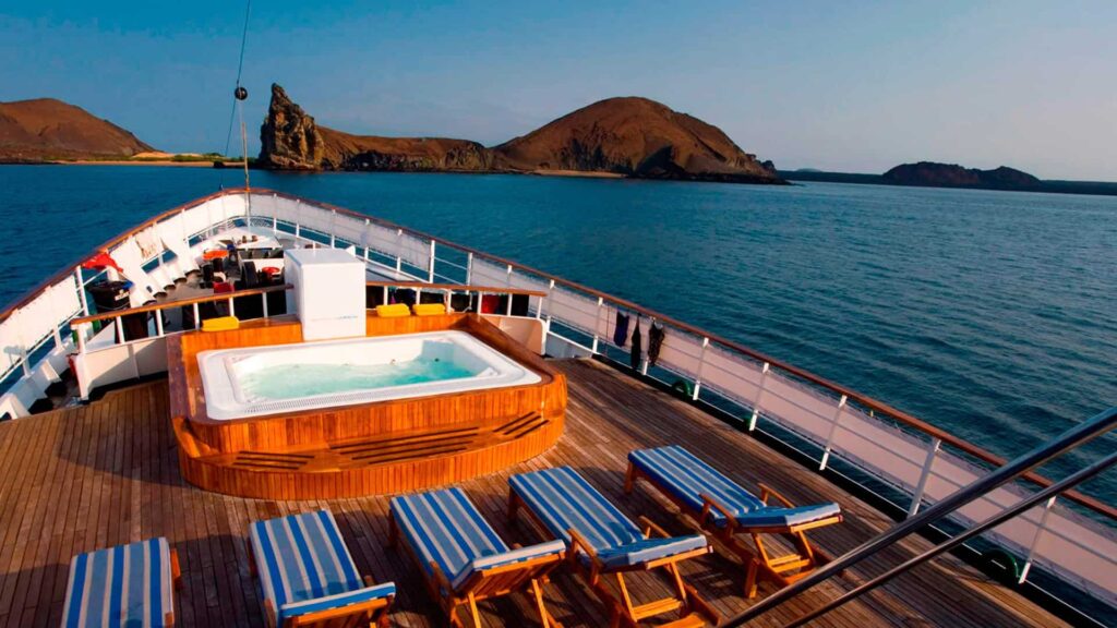Ship deck featuring a hot tub and several striped lounge chairs, overlooking calm blue waters and distant rocky islands under a clear sky.