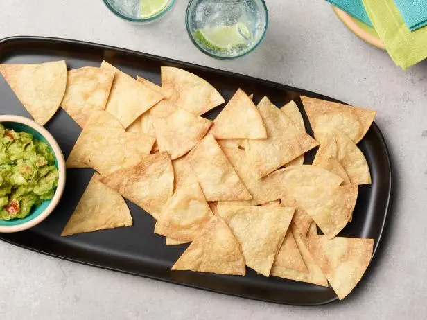 A black platter with crispy tortilla chips next to a bowl of guacamole. Two glasses with lime slices are partially visible in the background.