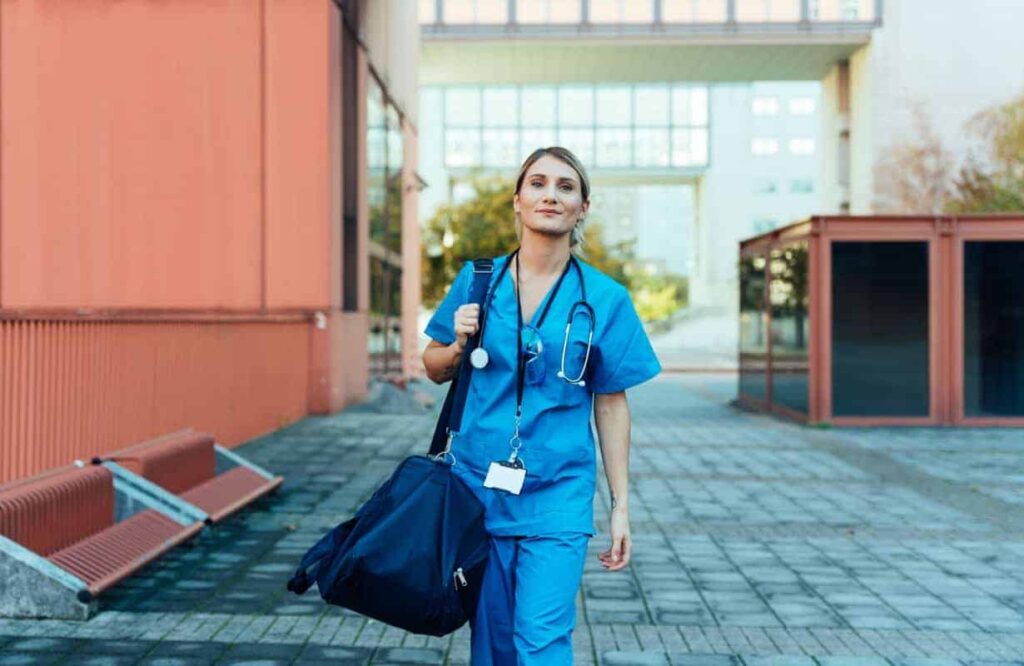 A healthcare professional in blue scrubs walks outdoors, carrying a medical bag and stethoscope, with buildings in the background.