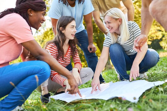 Group of people crouching on grass, engaged in discussion and pointing at a paper map spread out in front of them. They are outdoors in a park-like setting.