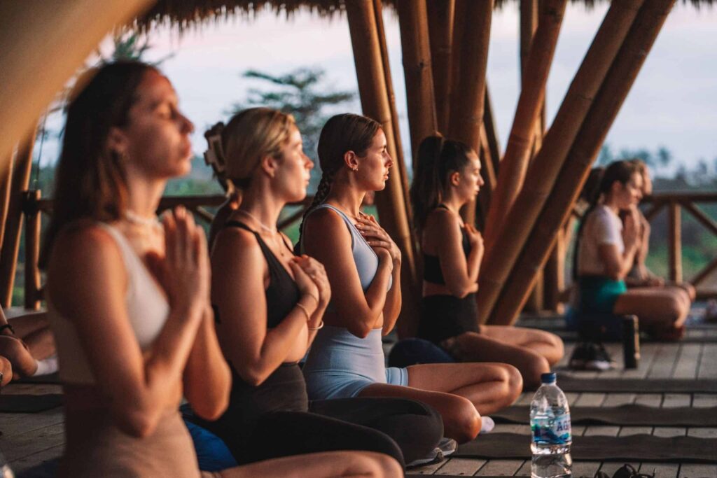 A group of people sit cross-legged on mats in a wooden pavilion, hands in prayer position, during a meditation or yoga session. A water bottle is visible in the foreground.