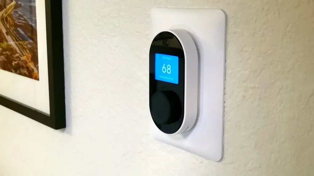 A smart thermostat is mounted on a wall, displaying a temperature of 68 degrees. A framed picture is partially visible beside it.