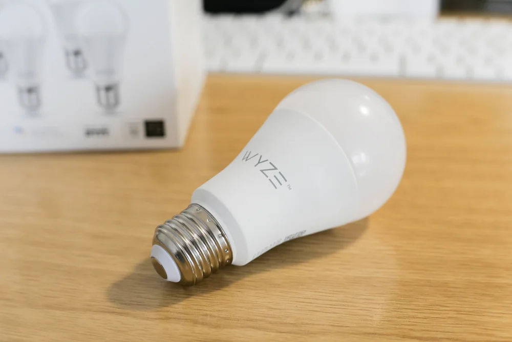 A white Wyze smart LED light bulb rests on a wooden surface, with its box partially visible in the background.