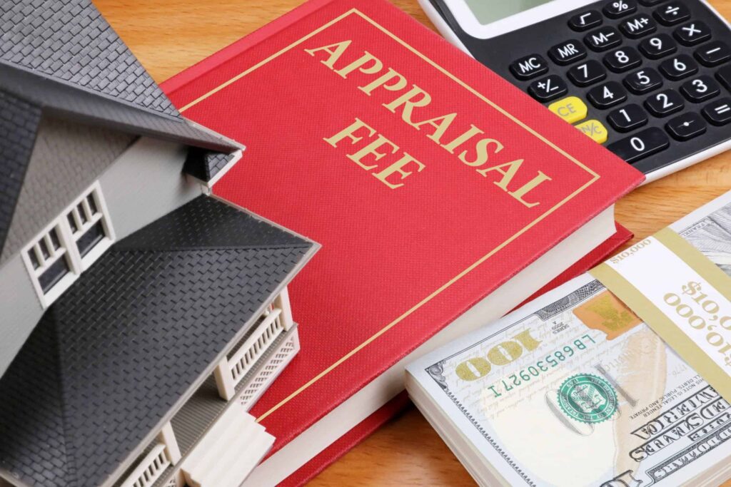 A model house, a red book labeled "APPRAISAL FEE," a calculator, and a stack of 100-dollar bills on a wooden surface.