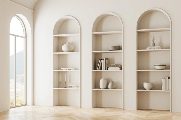 Minimalist room with three built-in arched shelves holding books, vases, and decorations. Large arched window on the left with sunlight streaming in.