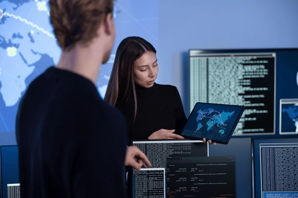 Two people in a control room analyzing data on screens and a tablet, showing maps and code.